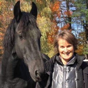 Joanne smiles at the camera wearing a shiny light purple jacket and standing beside a black horse